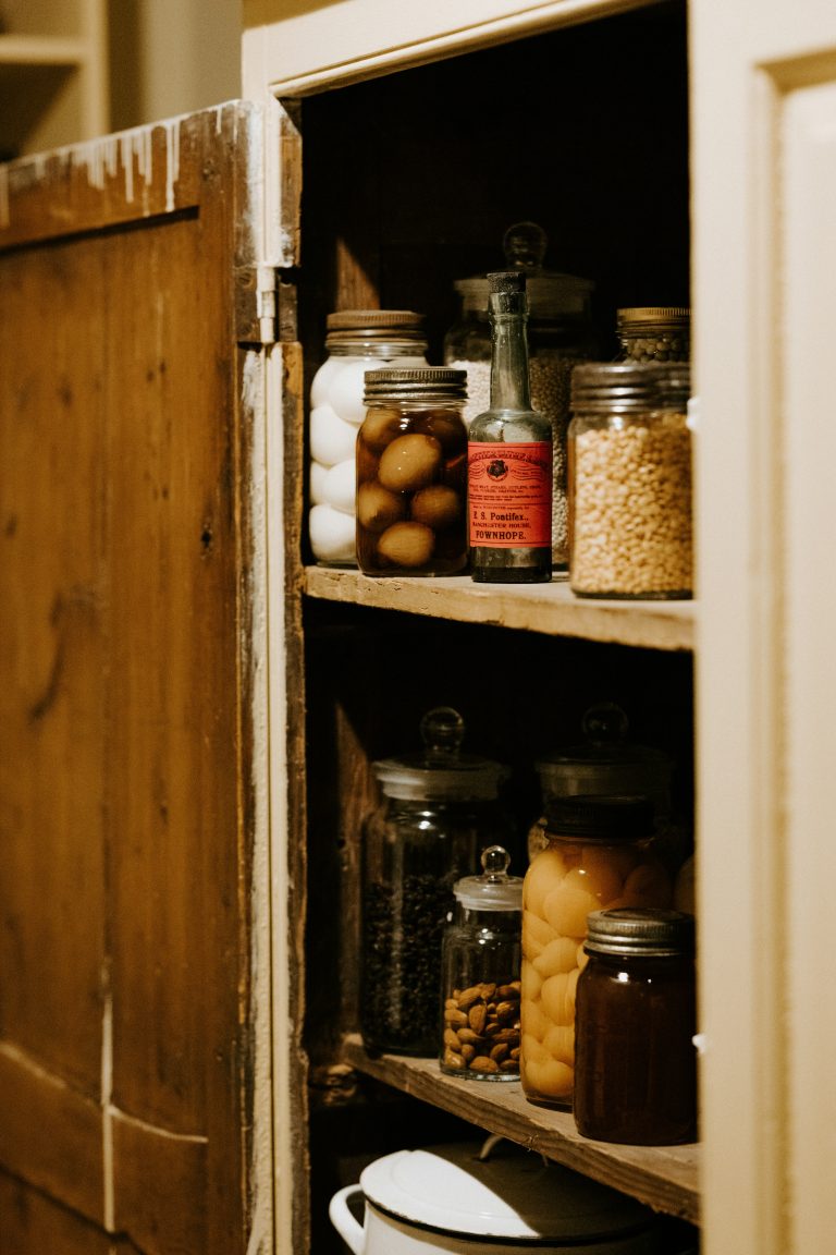 Open rustic pantry cupboard with wooden shelves stocked full of glass jars of preserved food, in warm dim light