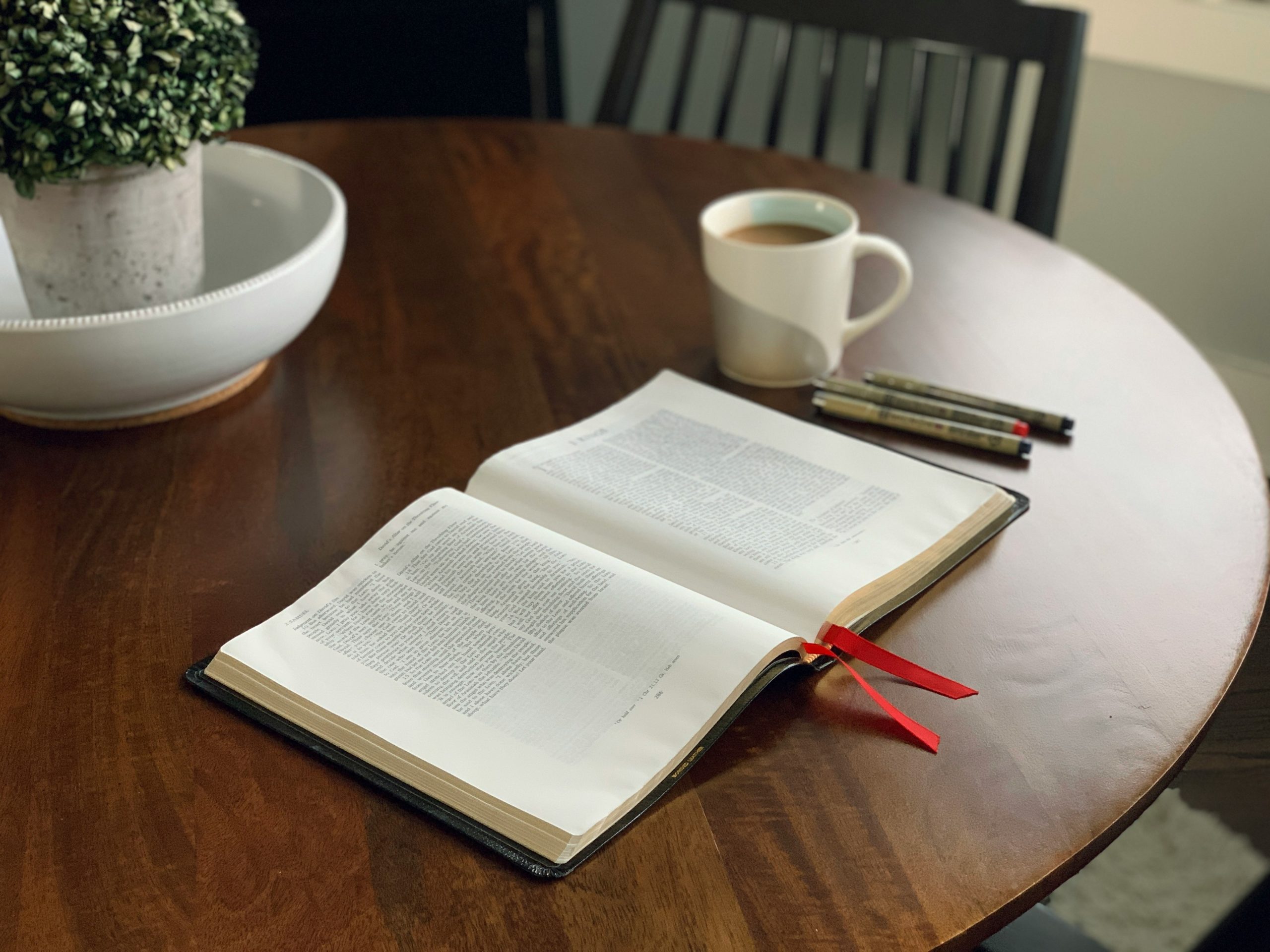 Open Bible with a red ribbon marker on a round wooden table, with a mug of coffee, pens and a chair