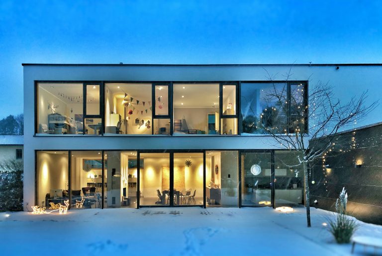 Modern house at dusk with floor-to-ceiling windows, every room lit and visible from outside